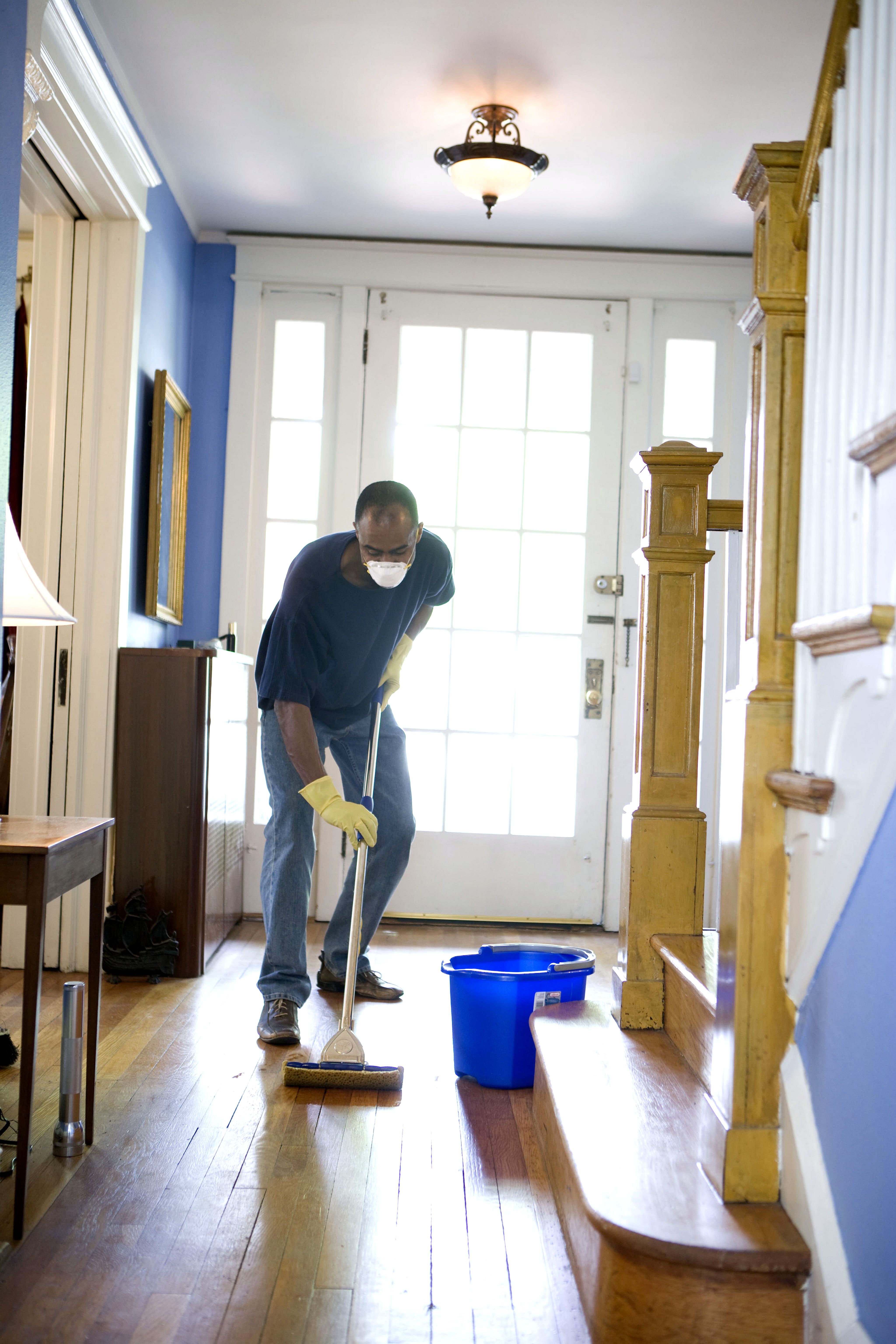 Cleaner mopping a hallway floor, wearing gloves and a dust mask, with a bucket beside them.